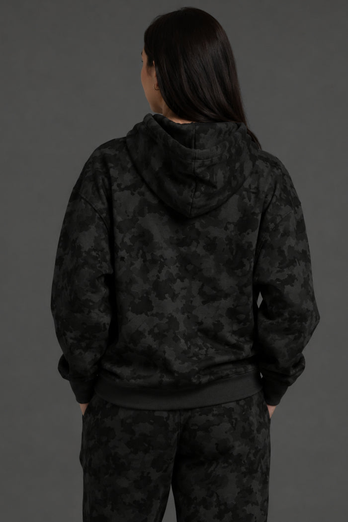 A person shown from the back wearing a black hooded sweatshirt and matching black patterned pants against a dark gray studio background. || Black Camo
