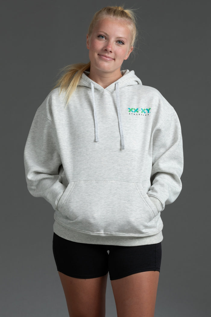A woman modeling a light heather gray pullover hoodie with a green 'XXX YYYathletics' logo on the left chest, paired with black athletic shorts, against a solid dark gray studio background. || Light Heather Gray