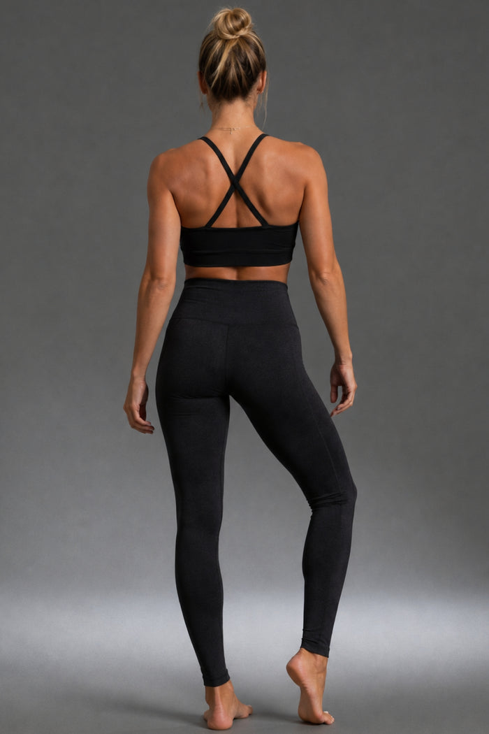 A woman is shown from behind wearing a black sports bra with crisscross back straps and black high-waisted leggings, standing against a neutral gray studio background. || Black Heather
